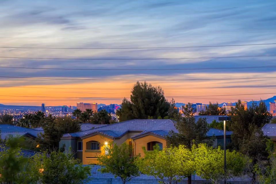 Sunset view over residential rooftops and trees with a distant illuminated city skyline.