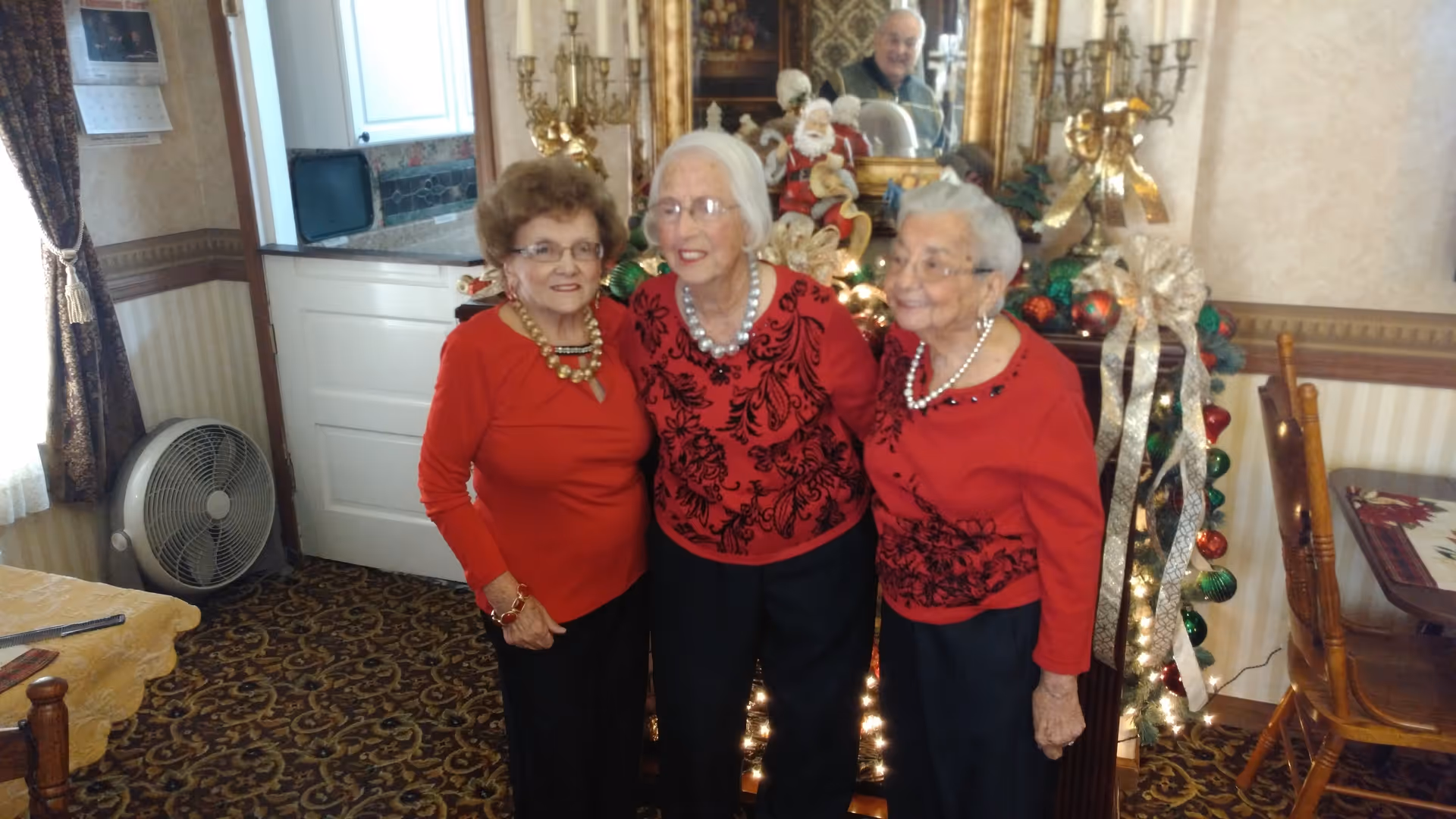 Three elderly women wearing red tops stand arm in arm in a festively decorated room with a mirror and holiday ornaments.