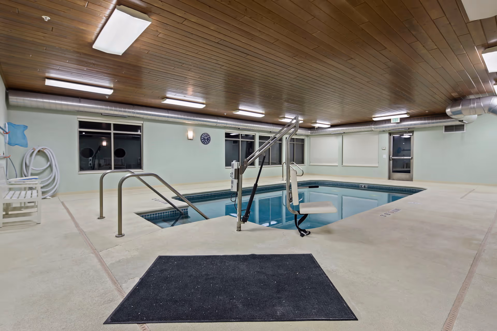 Indoor swimming pool with a mechanical lift, metal handrails, and bench seating under a wood-paneled ceiling.