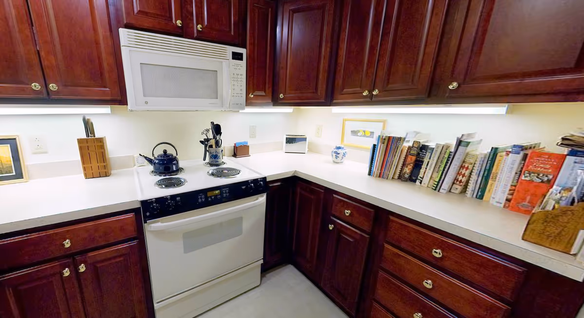 A kitchen corner with dark wooden cabinets and white countertops. There is a white electric stove with a blue kettle on it, a white microwave above the stove, a knife block, cooking utensils in a ceramic holder, and a collection of cookbooks lined up on the right side of the counter. The kitchen is well-lit with under-cabinet lighting.