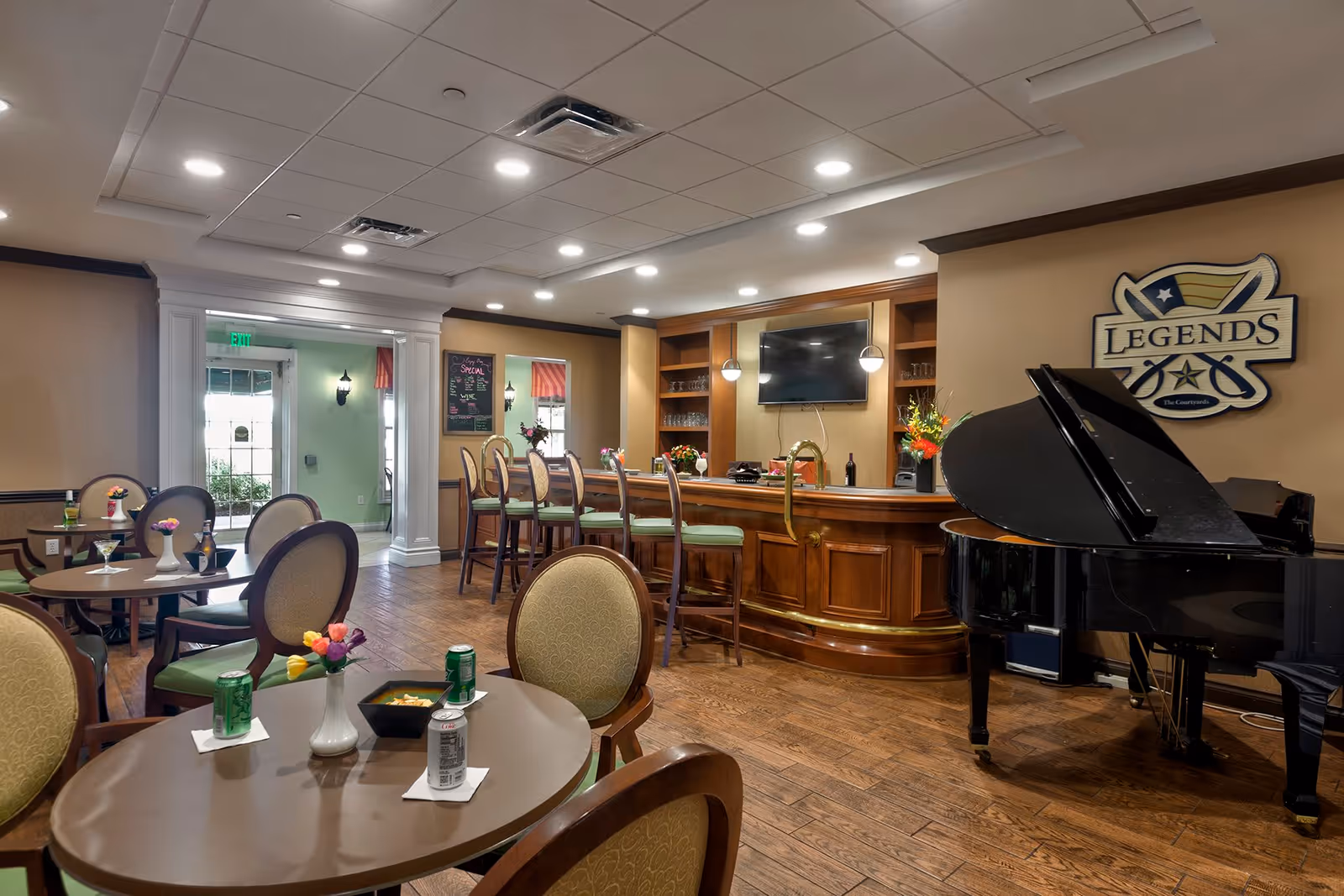 Interior view of a senior living facility lounge area with round tables and chairs, a wooden bar with high stools, a black grand piano, and a wall-mounted TV. The room has wooden flooring, recessed ceiling lights, and a sign on the wall that reads 'Legends The Courtyards'.