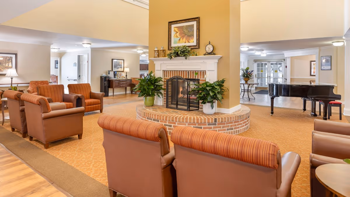 A senior living facility lounge with armchairs arranged around a central brick fireplace and a grand piano in the background.