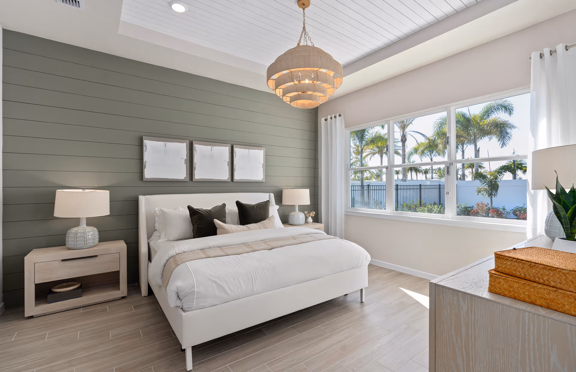 A bright and modern bedroom with a large white bed featuring white and dark pillows, two bedside tables with lamps, a green shiplap accent wall with three abstract paintings above the bed, a large window with white curtains showing palm trees outside, and a light wood dresser with decorative items.