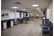 A spacious rehabilitation or physical therapy room with exercise equipment, chairs lined up along the walls, and large windows with blinds. The room has a light-colored wood floor and a white ceiling with recessed lighting.