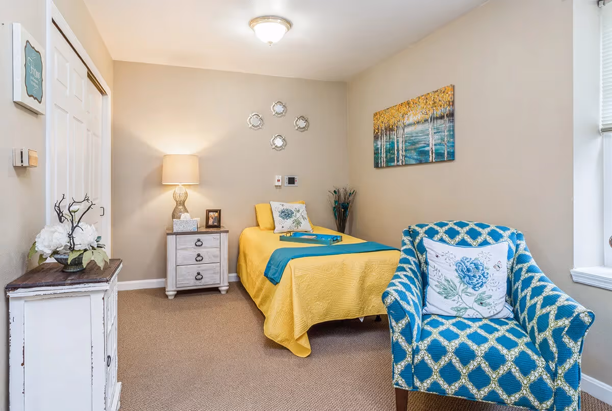 A cozy single-occupancy bedroom with a yellow bed, patterned blue armchair, nightstand with lamp, and wall art.