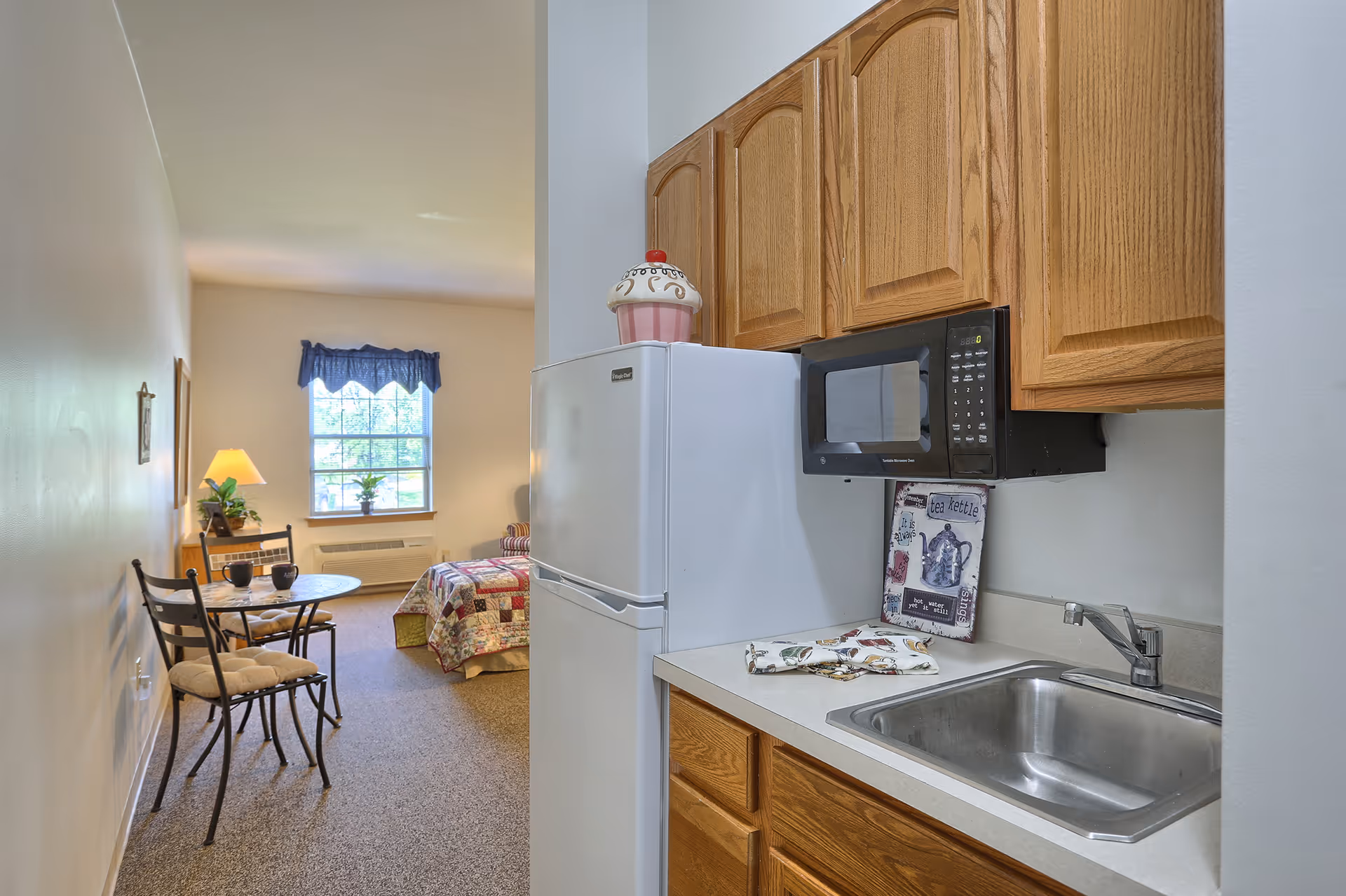 A small kitchen area with wooden cabinets, a white refrigerator, a microwave, and a stainless steel sink. Beyond the kitchen, there is a small dining table with two chairs, a lamp on a side table, a window with a blue valance, and a bed with a colorful quilt in a cozy living space.