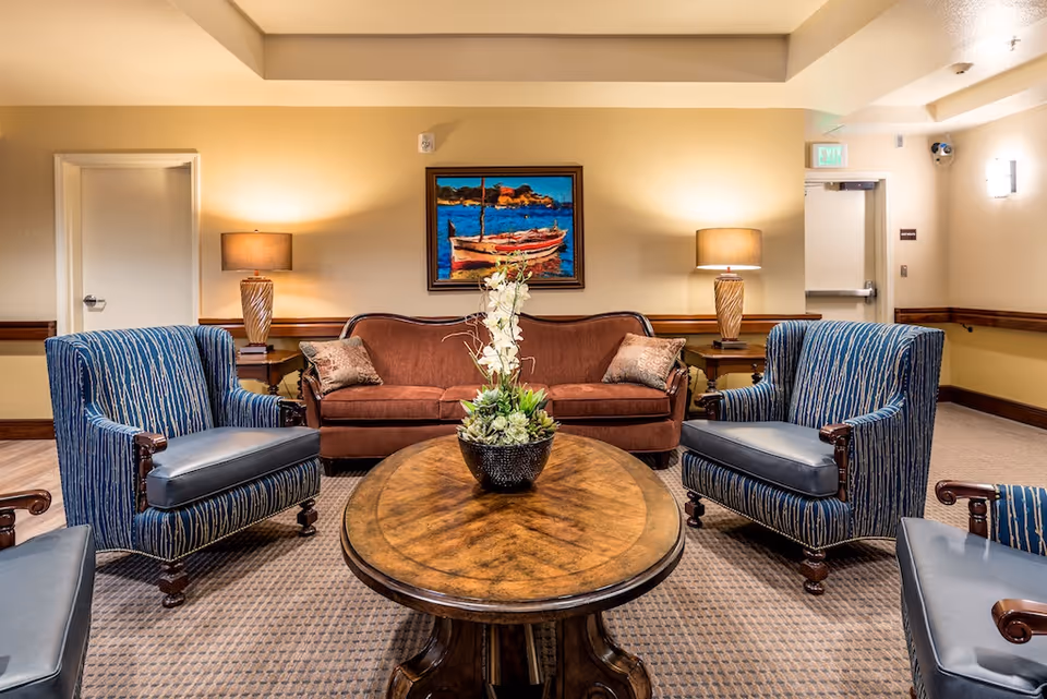A cozy senior living facility lounge area with a brown sofa, two blue patterned armchairs, a round wooden coffee table with a floral centerpiece, two table lamps on side tables, and a colorful painting of a boat on the wall.