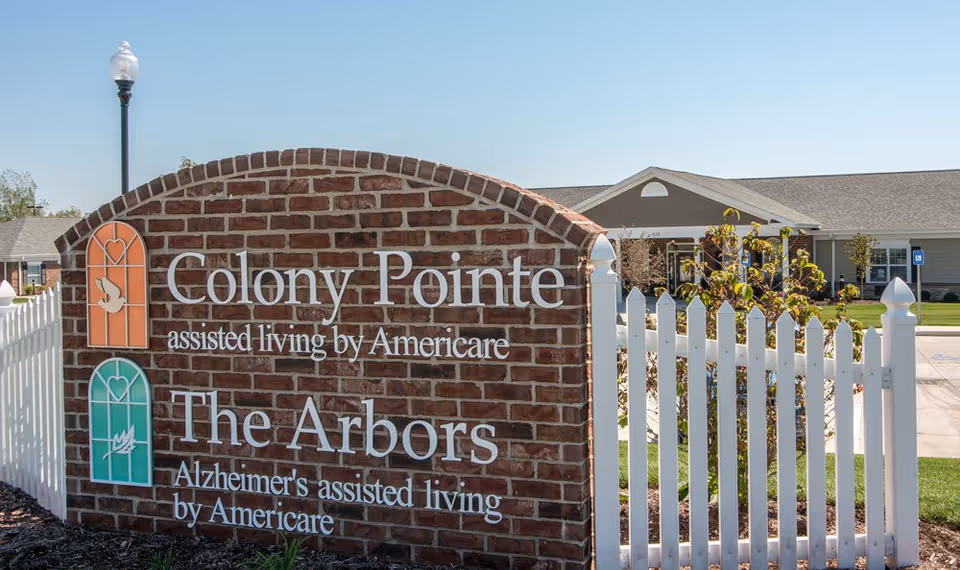 Brick sign at the entrance of Colony Pointe Senior Living and The Arbors Alzheimer's assisted living by Americare, with a white picket fence and a building in the background under a clear blue sky.