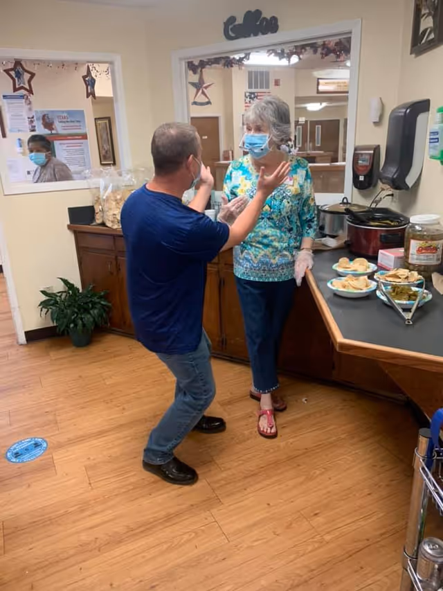 A man and a woman wearing face masks are engaged in conversation in a room with wooden flooring and a counter with plates of food. The woman is standing near the counter, and the man is gesturing with his hands. There are decorations on the walls and a window showing another person wearing a mask.