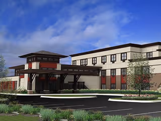 Exterior view of a modern three-story healthcare facility building with a covered entrance, surrounded by landscaped greenery and a parking area under a partly cloudy blue sky.