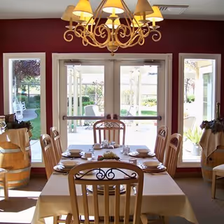 Dining room with a rectangular table set for six people with plates, bowls, and cups. The room has a burgundy wall with large glass doors and windows looking out onto a garden patio. There is a chandelier hanging above the table and decorative barrels with plants on either side of the doors.