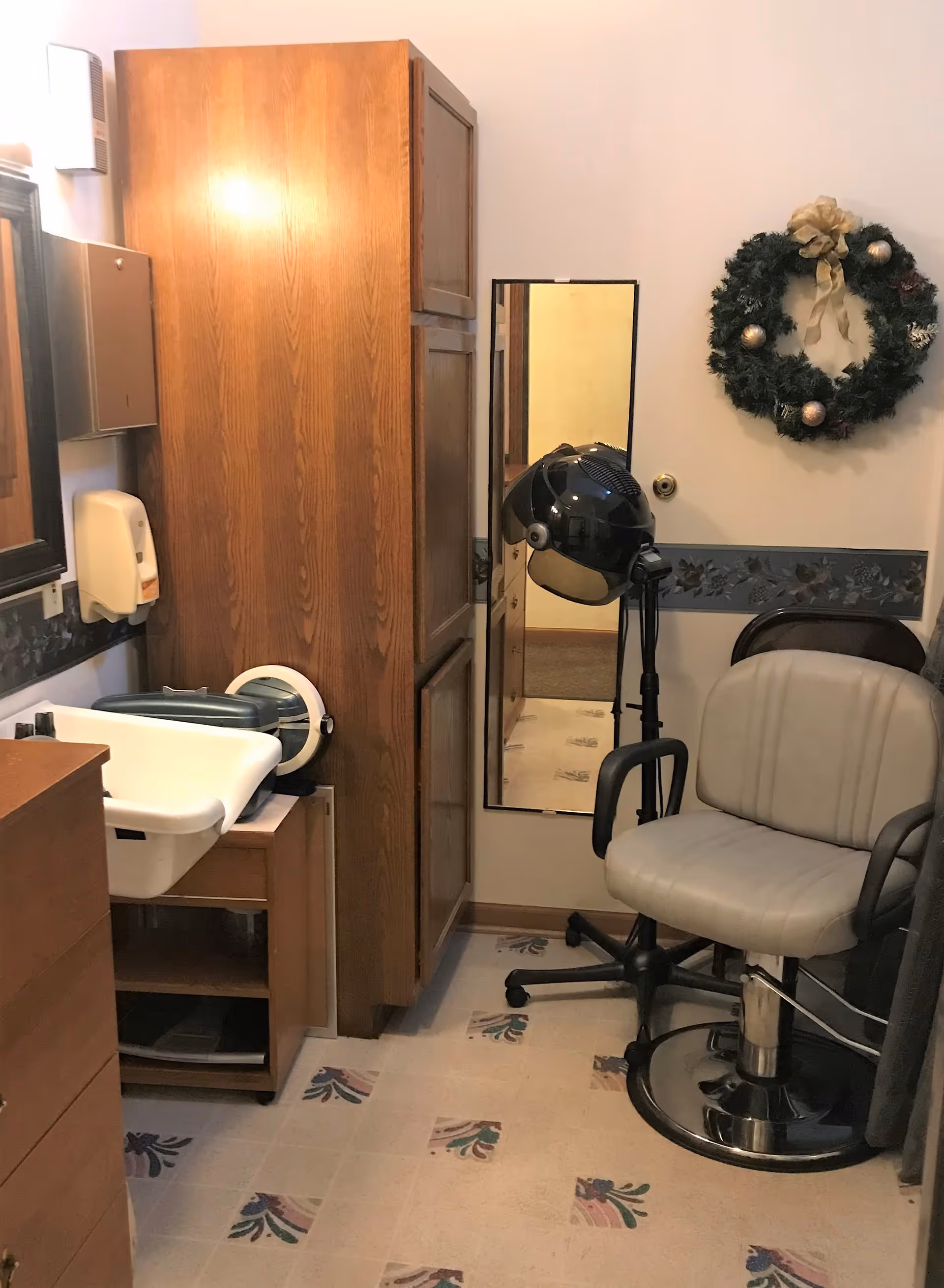 A small salon or hair styling area with a beige salon chair, a black hair dryer on a stand, a tall wooden cabinet, a wall-mounted mirror, a sink, and a decorative wreath hanging on the wall.
