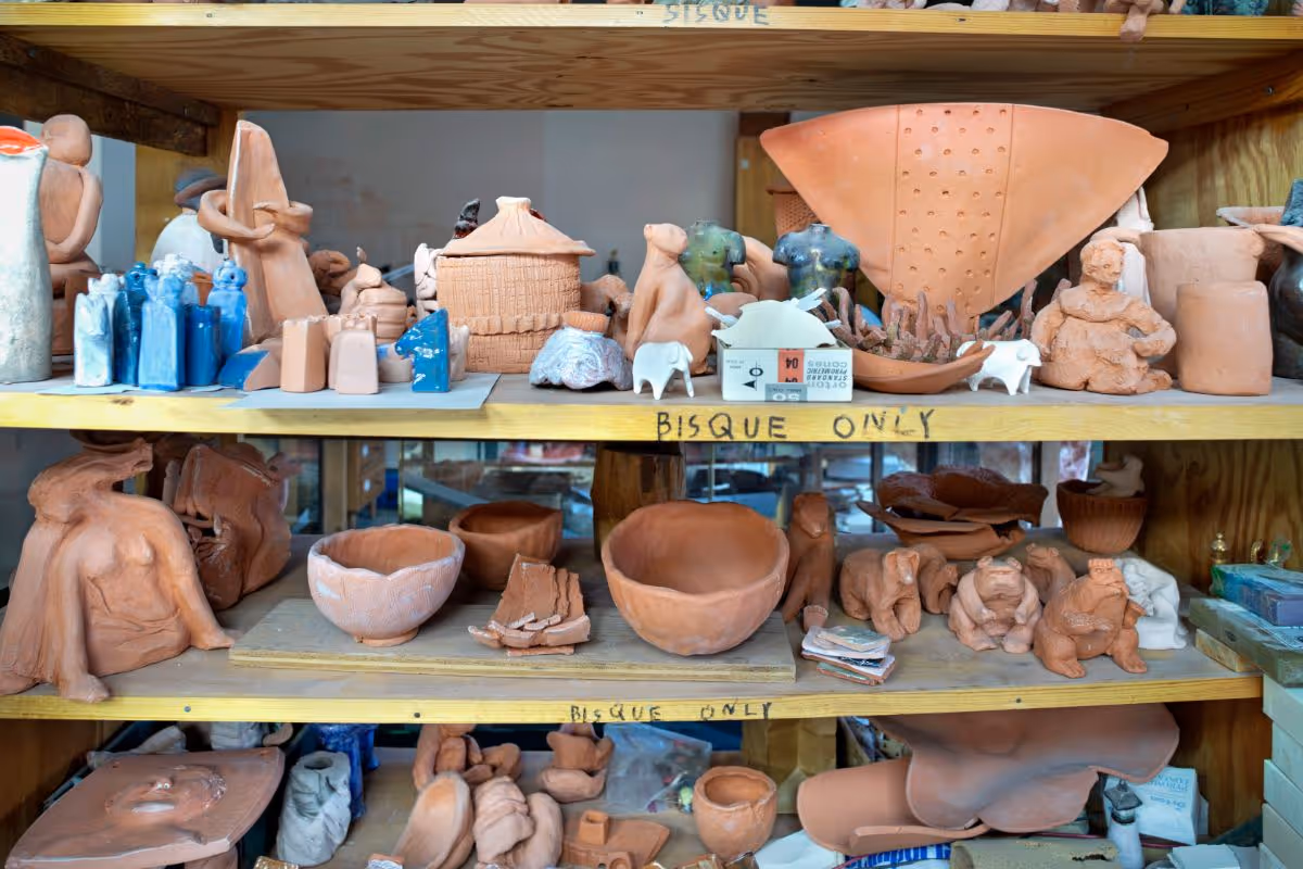 Wooden shelves filled with various bisque pottery items including bowls, figurines, and small sculptures in an art studio or workshop setting.