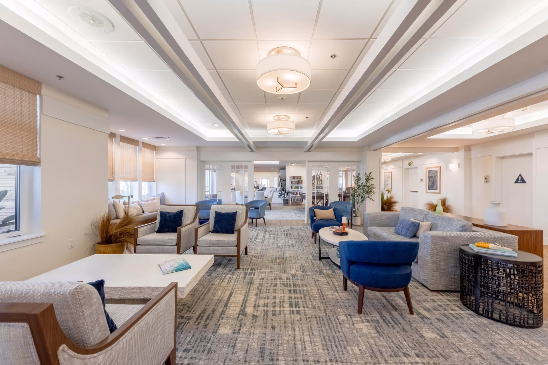 A spacious and well-lit common area in a senior living facility featuring modern furniture including beige armchairs with dark blue cushions, a gray sofa, and blue chairs arranged around white and black coffee tables. The room has large windows with woven shades, a patterned carpet, and ceiling lights. In the background, there is a glimpse of a bookshelf and additional seating areas.