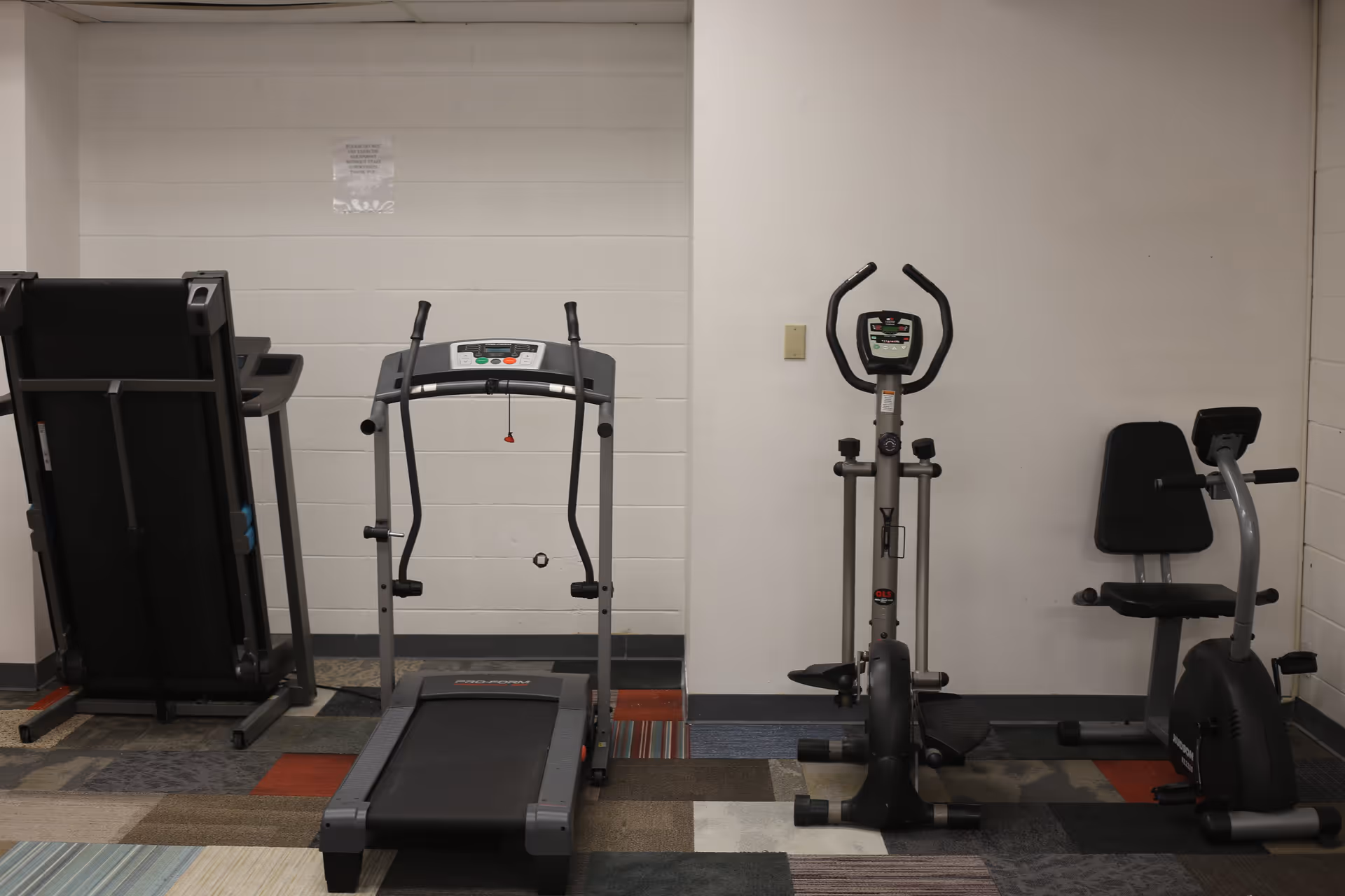 Indoor exercise room with a treadmill, elliptical machine, and stationary bike against a white wall with a multicolored carpeted floor.