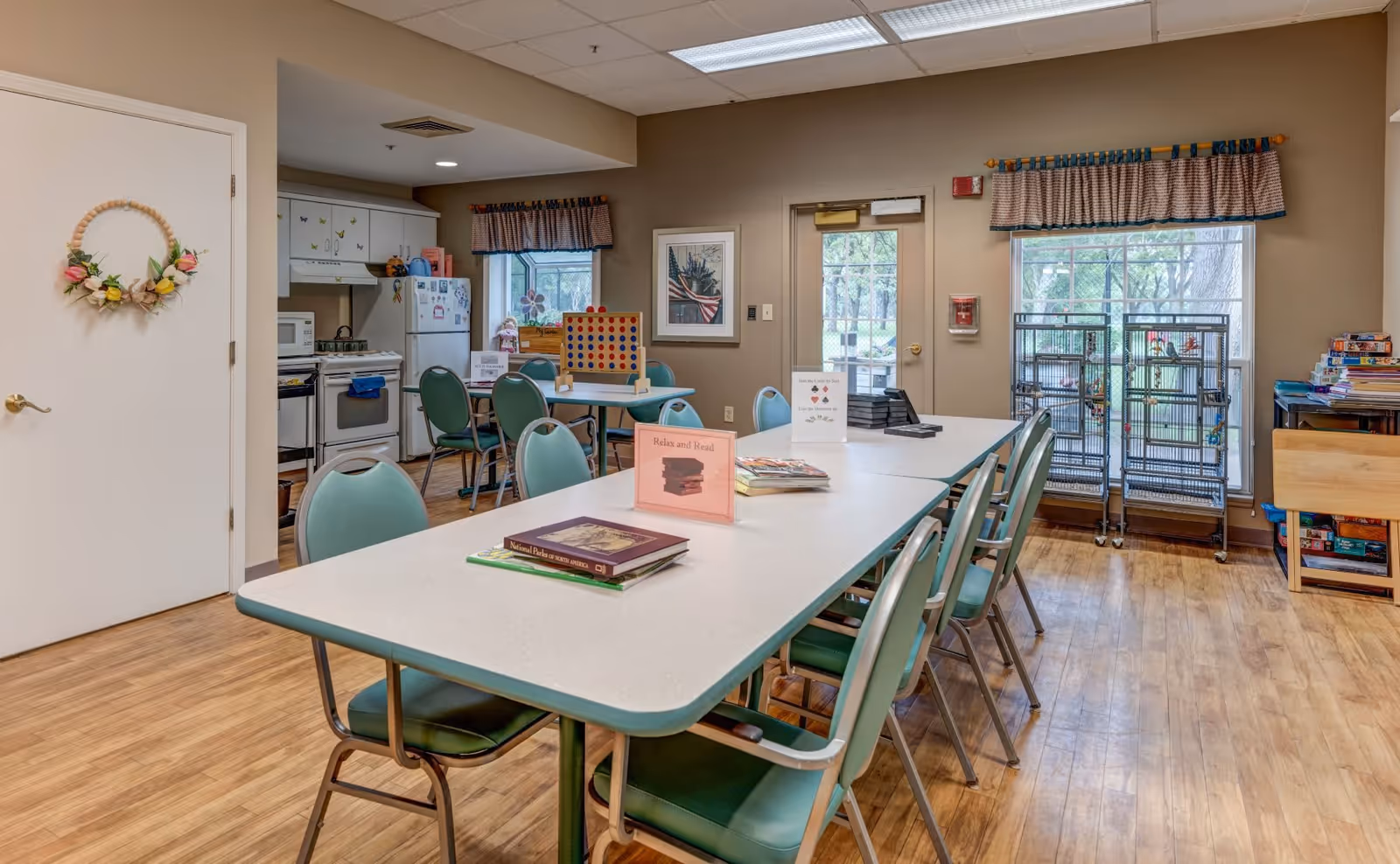 A communal room with a long white table surrounded by green chairs. On the table are books and signs, one reading 'Relax and Read.' In the background, there is a small kitchen area with a refrigerator, stove, and microwave. A window with a valance and a door leading outside are visible, along with shelves holding board games and other items. The floor is wooden, and the walls are beige.