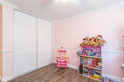 A small room with light pink walls and white trim, featuring a white double-door closet on the left. On the right side, there is a pink toy kitchen set and a dollhouse filled with various toys and stuffed animals on top. The floor is a medium-tone wood laminate.