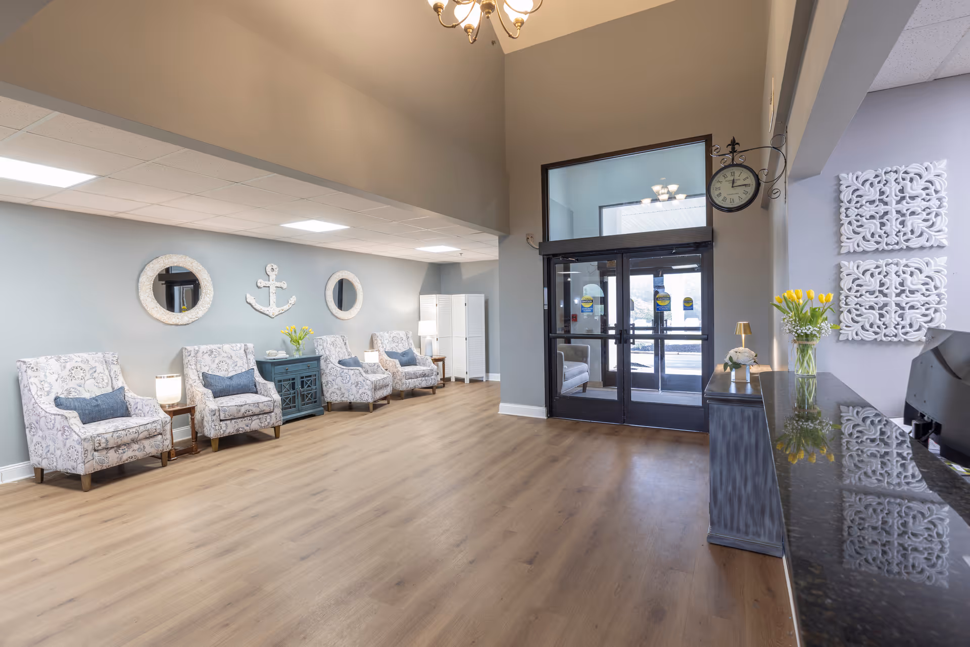 A spacious and well-lit assisted living facility lobby with light wood flooring, a row of patterned armchairs with blue cushions along a gray wall decorated with two round mirrors and an anchor wall art. There is a black granite reception desk on the right with a vase of yellow tulips and decorative items. Double glass entrance doors are visible at the far end, with a wall clock mounted above them. The ceiling is high with a chandelier hanging down.