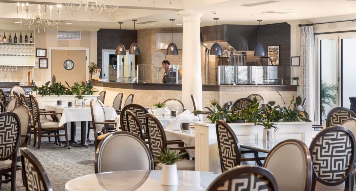 A bright and elegant dining room with multiple tables covered in white tablecloths, surrounded by chairs with patterned upholstery. The room features a kitchen area in the background with hanging pendant lights and a person working behind the counter. There are plants placed on dividers between tables and large windows with curtains allowing natural light to enter.