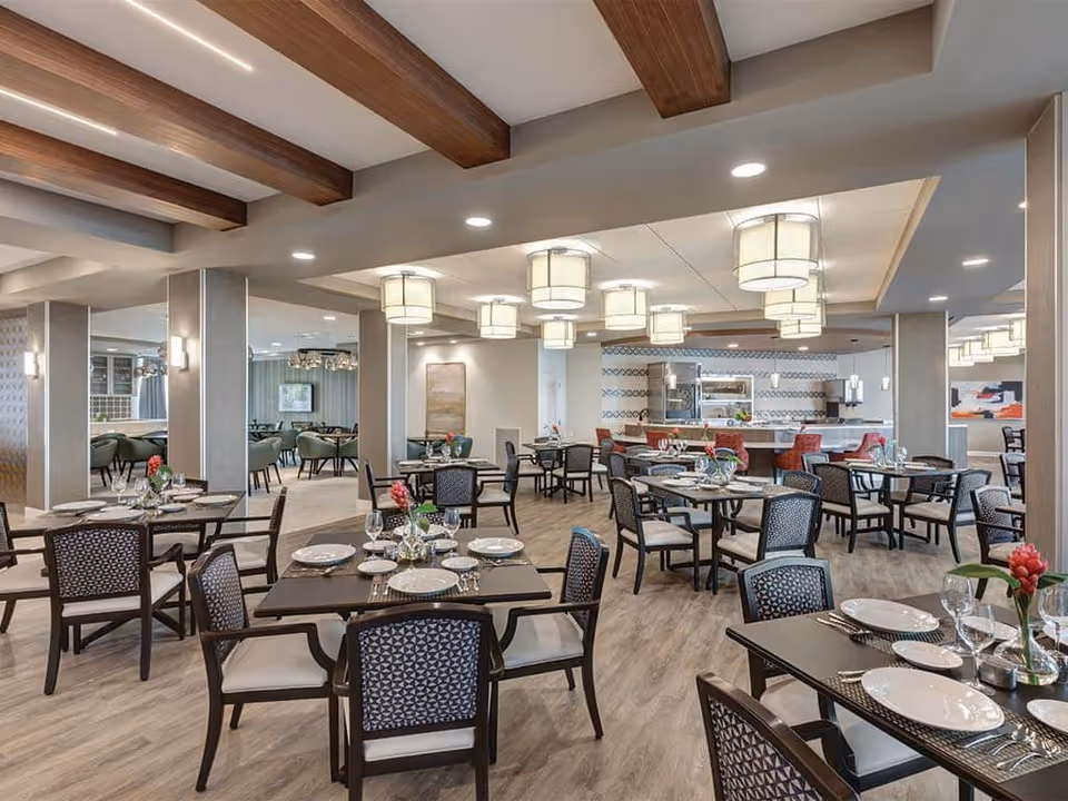 A spacious and well-lit dining room with multiple tables set with plates, glasses, and silverware. The room features wooden beams on the ceiling, modern hanging light fixtures, and a mix of dark wood and cushioned chairs. There are decorative flowers on each table and a kitchen area visible in the background with red chairs at a counter.