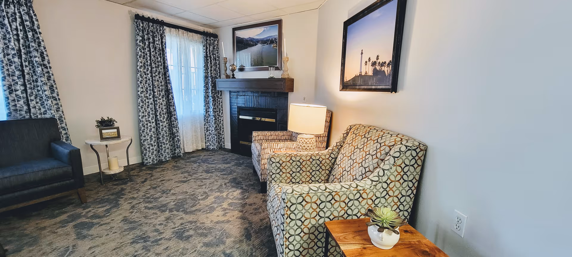 A cozy living room area with patterned armchairs, a dark blue sofa, a small wooden side table with a potted plant, a lamp, and a fireplace with decorative items on the mantel. The room has patterned curtains and framed landscape photographs on the walls.