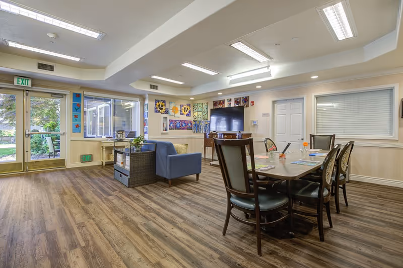 A bright and spacious common area in a senior living facility with wooden flooring, a table with six chairs, a blue armchair, a TV mounted on a wooden cabinet, colorful artwork on the walls, and large windows and glass doors leading to an outdoor garden area.