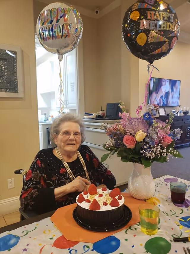 An elderly woman sitting at a table with a birthday cake topped with strawberries and cherries in front of her. Two 'Happy Birthday' balloons are floating above her. A vase with a colorful bouquet of flowers is on the table, which is covered with a festive tablecloth. In the background, a television is on and some furniture is visible.