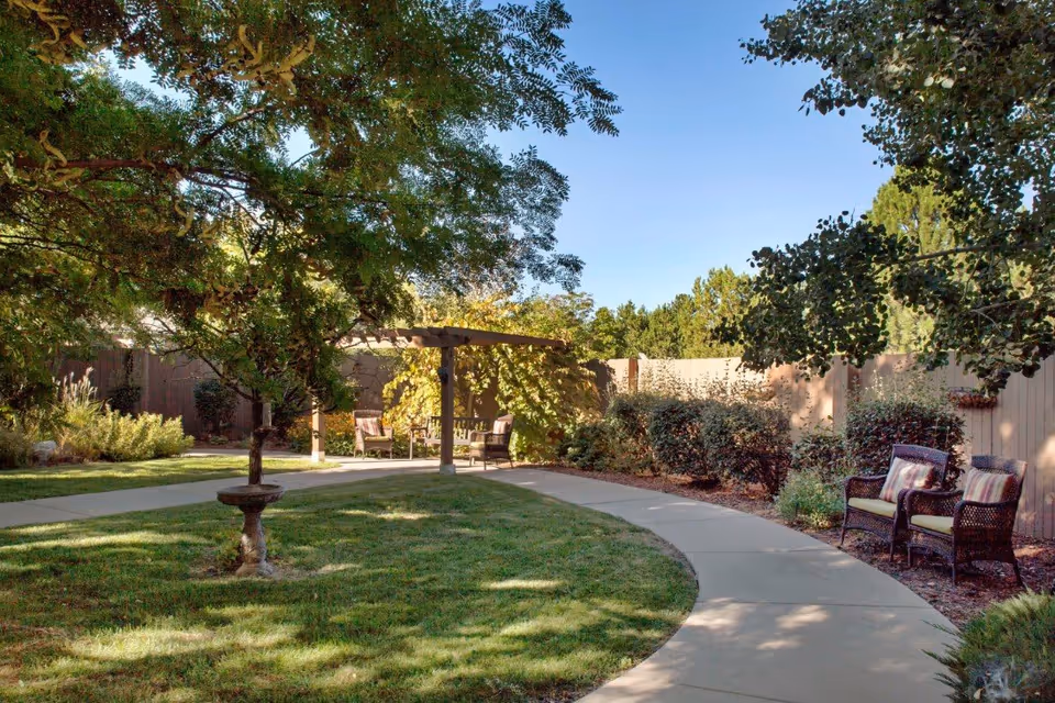 A peaceful outdoor garden area with a curved concrete pathway surrounded by green grass, trees, and bushes. There are two wicker chairs with cushions on the right side and a wooden pergola with additional seating in the background. The sky is clear and blue.