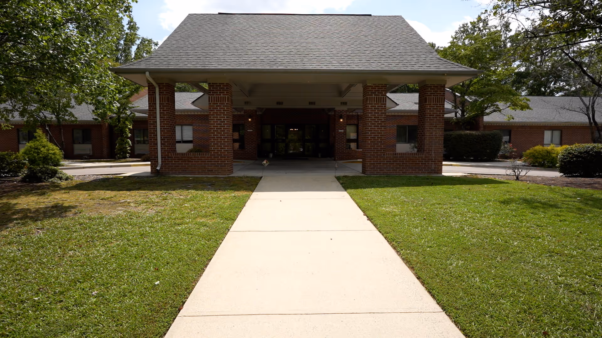 Front entrance of a single-story brick building with a covered walkway supported by brick columns, surrounded by green grass and trees under a partly cloudy sky.