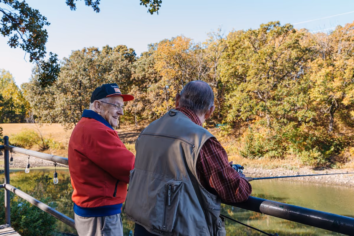 Two elderly men fishing off a wooden railing by a calm pond surrounded by trees with autumn foliage under a clear blue sky.