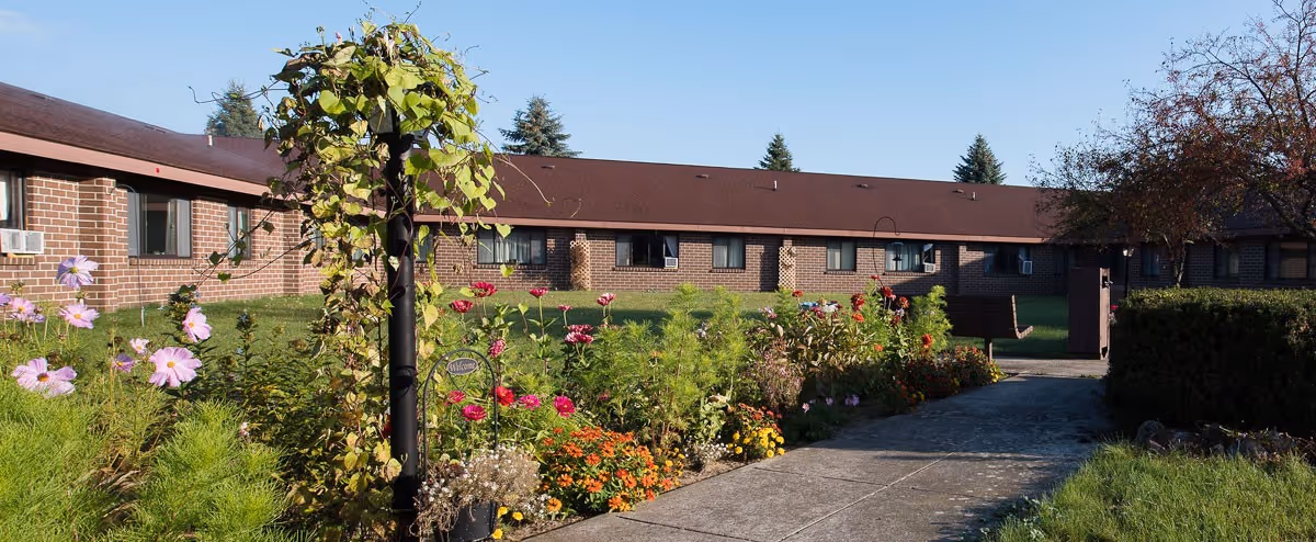 Outdoor garden area with colorful flowers and greenery along a paved walkway leading to a brick building with multiple windows and a brown roof under a clear blue sky.