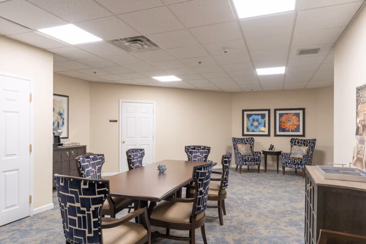 A well-lit interior room with a rectangular wooden table surrounded by six patterned chairs. In the background, there are two armchairs with matching patterned upholstery and a small round table between them. The walls are beige with two framed floral artworks hanging above the armchairs. There is a cabinet with a coffee machine and cups on the left side, and a closed white door in the back wall. The carpet is blue with a subtle pattern, and the ceiling has recessed lighting panels.
