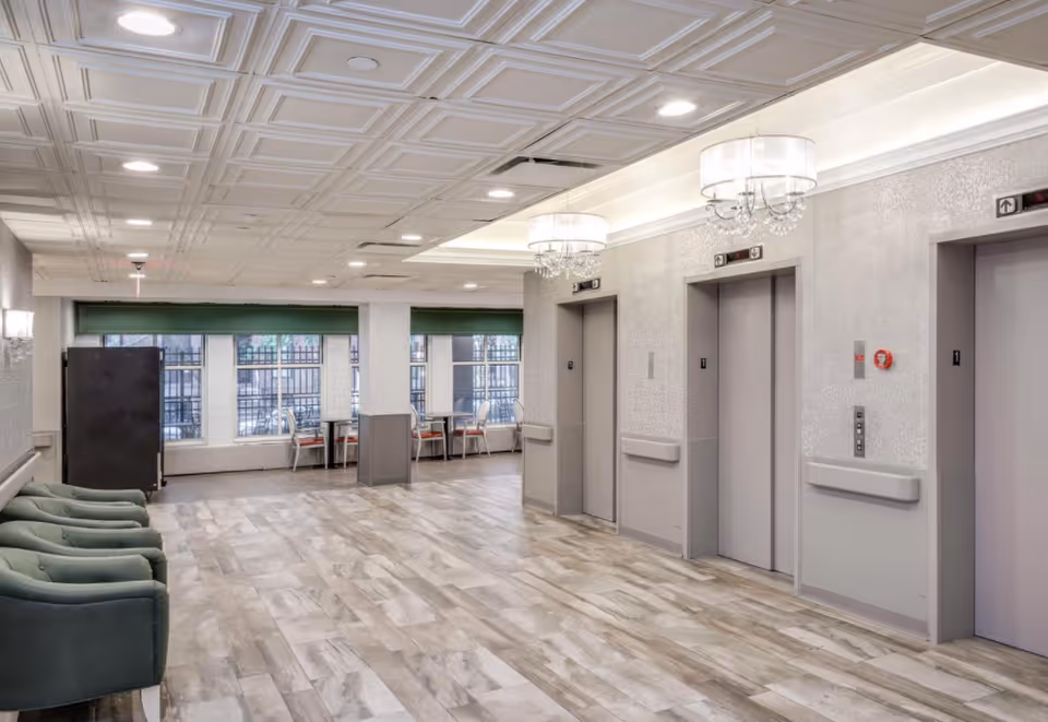 Interior view of a hallway area in a senior living facility with three closed elevators on the right side, green cushioned chairs on the left, and tables with chairs near large windows in the background. The ceiling has recessed lighting and decorative light fixtures.