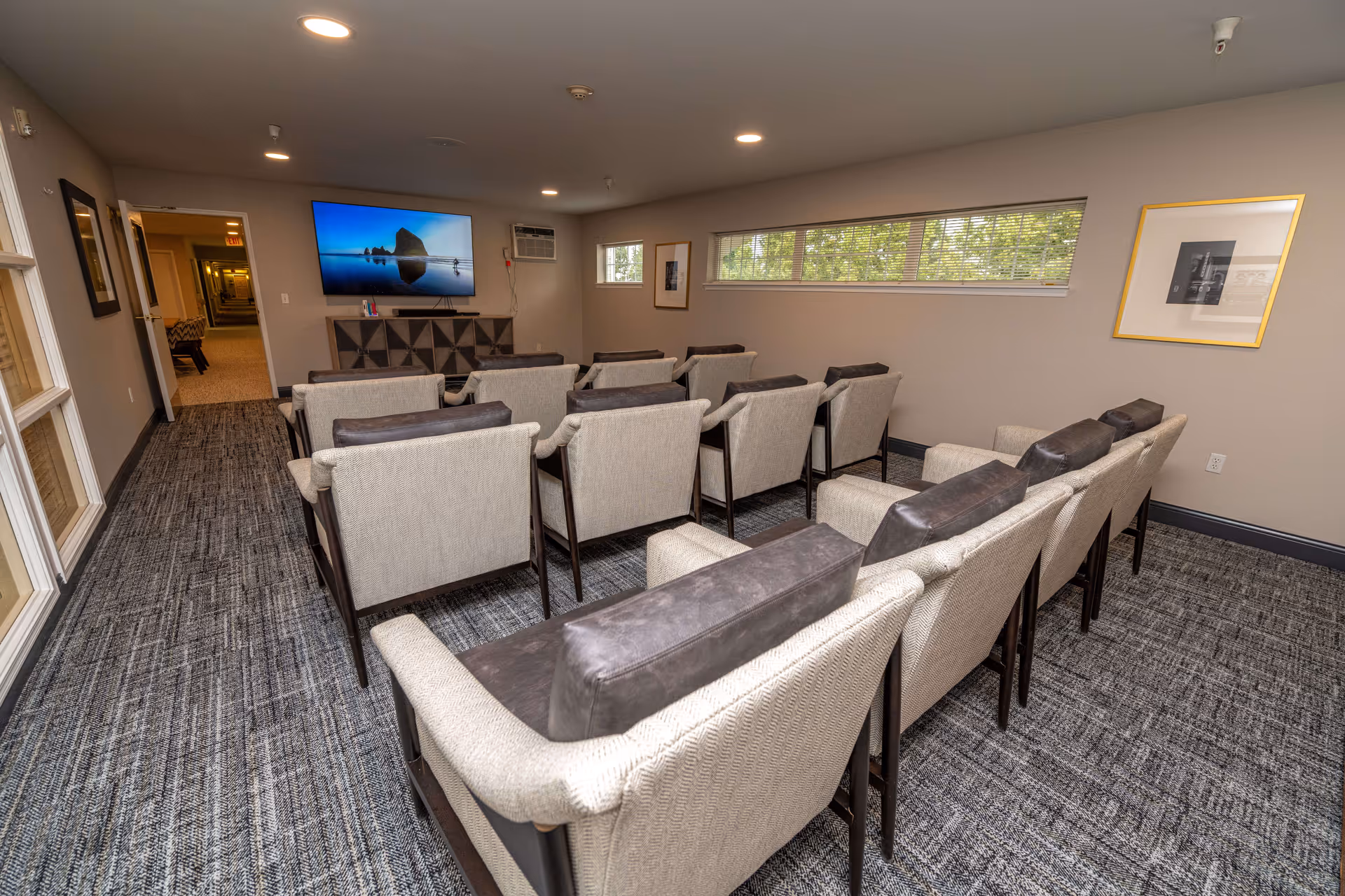 A small theater-style room with three rows of beige cushioned chairs facing a wall-mounted flat-screen TV. The room has gray carpet, beige walls, a long horizontal window with blinds, framed pictures on the walls, and recessed ceiling lights.