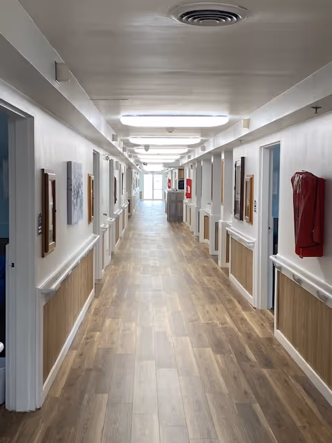 A long, well-lit hallway in a senior living facility with wooden flooring and white walls. The hallway has handrails on both sides and several doorways leading to rooms. There are framed pictures on the walls and fire safety equipment mounted on the right side.