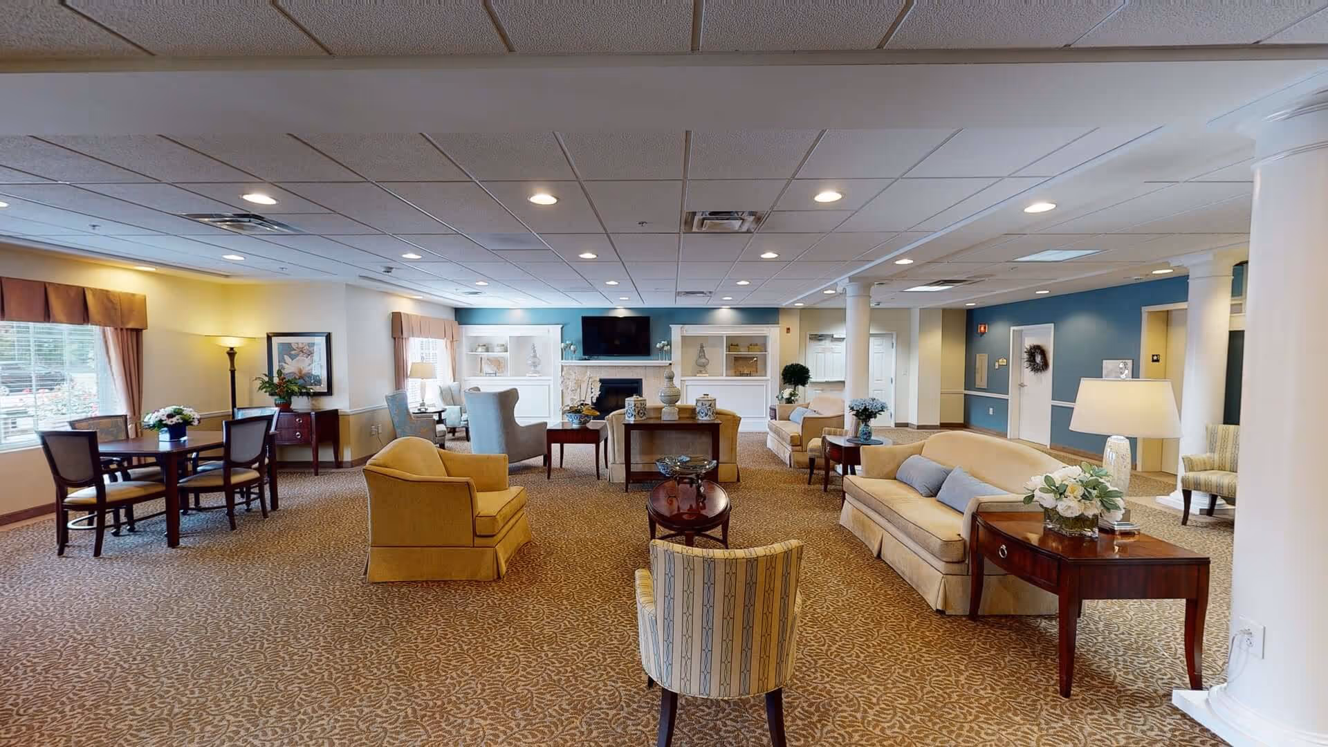 A spacious and well-lit living room area in a senior living facility with multiple seating arrangements including sofas, armchairs, and small tables. The room features carpeted flooring, a fireplace with a TV mounted above it, decorative shelves, and large windows with curtains allowing natural light to enter. The walls are painted in soft colors with some blue accent walls and white pillars.