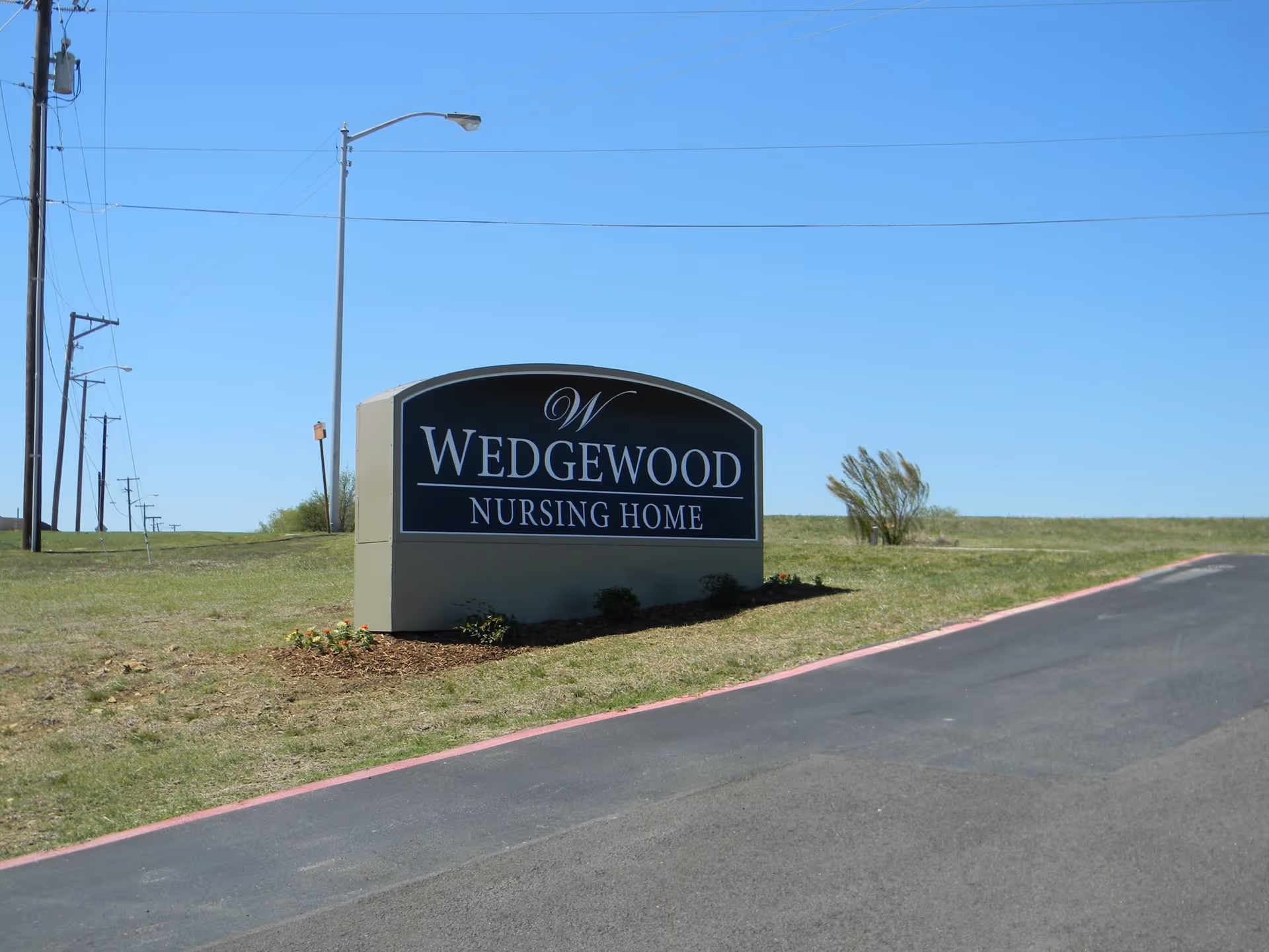 A large sign for Wedgewood Nursing Home situated on a grassy area next to a paved road under a clear blue sky.