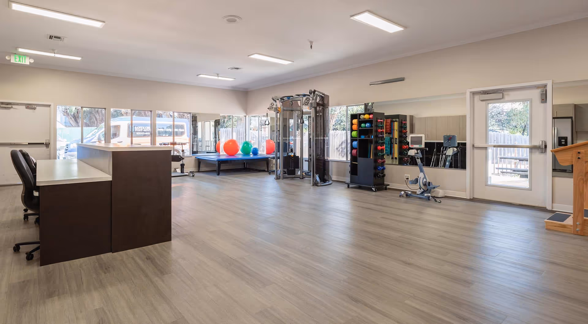 A spacious rehabilitation or physical therapy room with light wood flooring, a reception desk with chairs, exercise equipment including a multi-functional cable machine, colorful exercise balls on a raised platform, a rack with various weights and exercise mats, a stationary bike, and large mirrors on the walls. Large windows and a glass door allow natural light to enter the room.