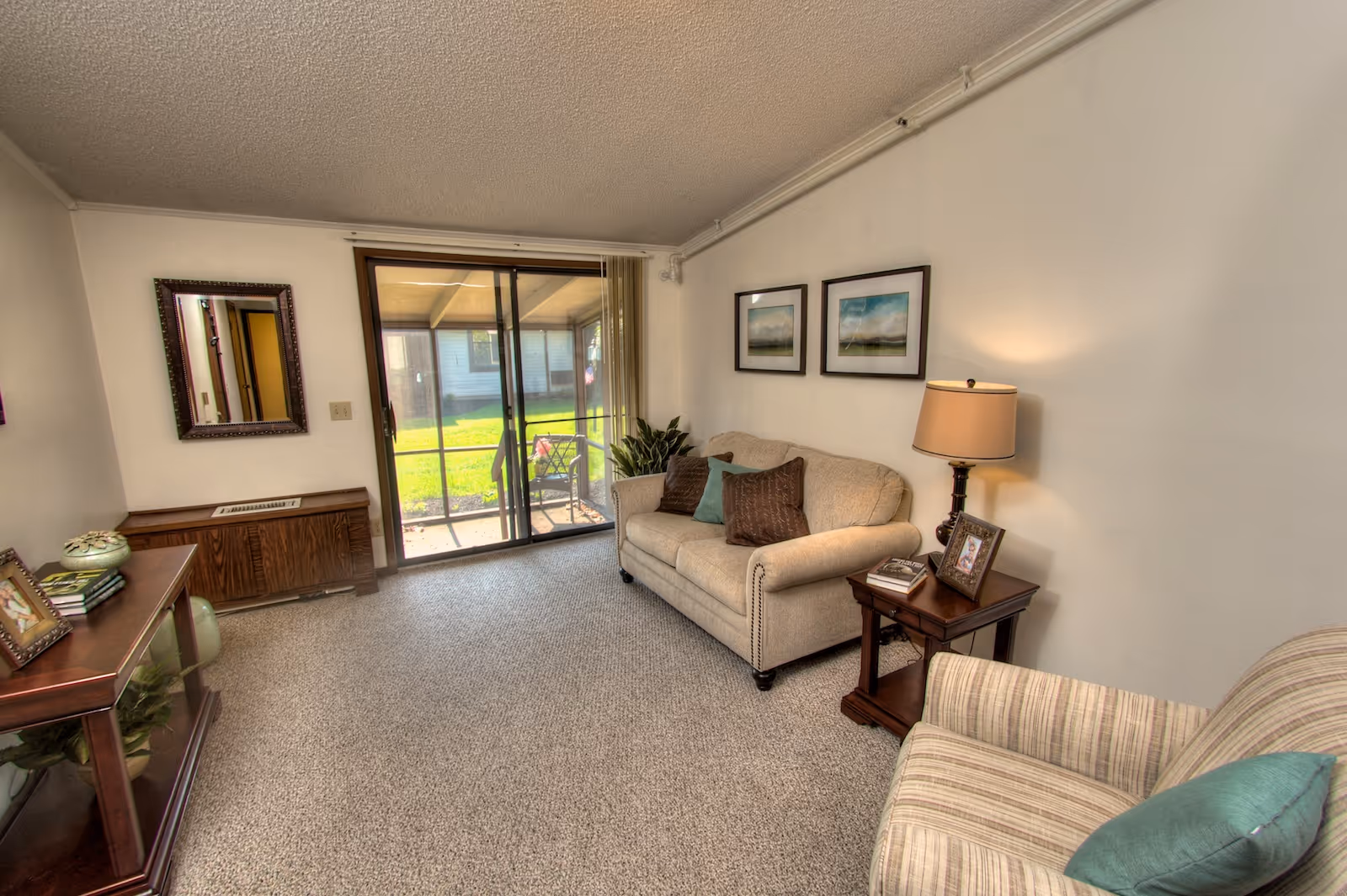 A cozy living room in Springfield Assisted Living featuring a beige sofa with decorative pillows, a striped armchair with a teal pillow, a wooden side table with a lamp and framed photo, a wooden console table with books and decorative items, a wall mirror, and sliding glass doors leading to a green outdoor area.