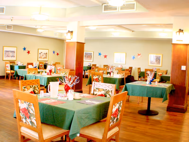 Dining room with multiple tables covered in green tablecloths and wooden chairs arranged for meals.