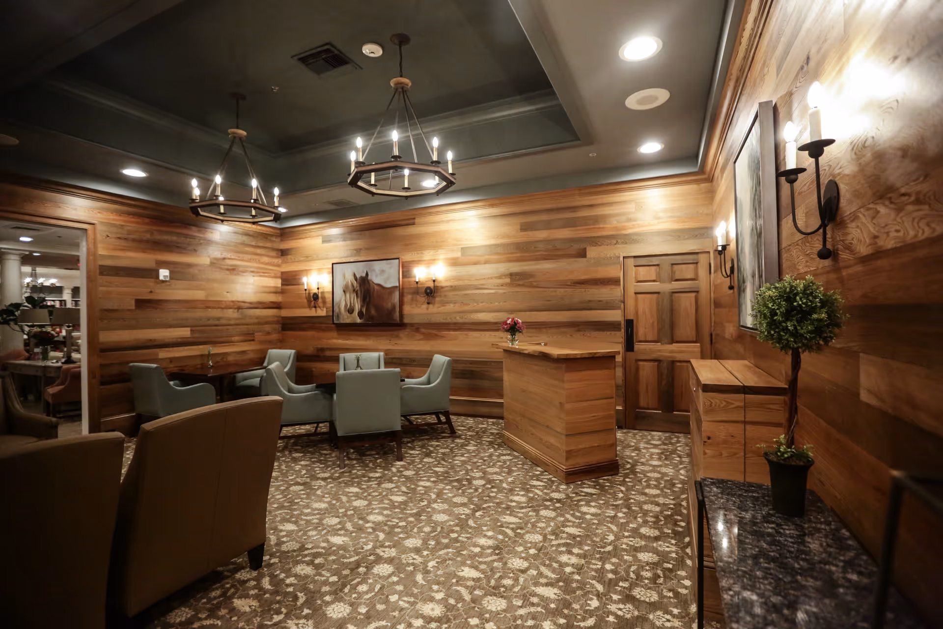 Wood-paneled lounge with chandeliers, armchairs grouped around a table and a small reception desk.