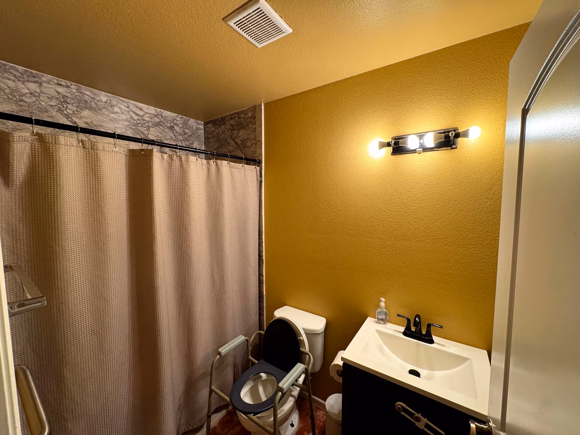 A small bathroom with a beige shower curtain, a toilet with a raised toilet seat and arm supports, a white sink with a black faucet, a soap dispenser, and a yellow wall with a light fixture above the sink.