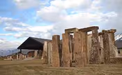 Stonehenge-inspired upright stones stand on a grassy field with a building in the background.