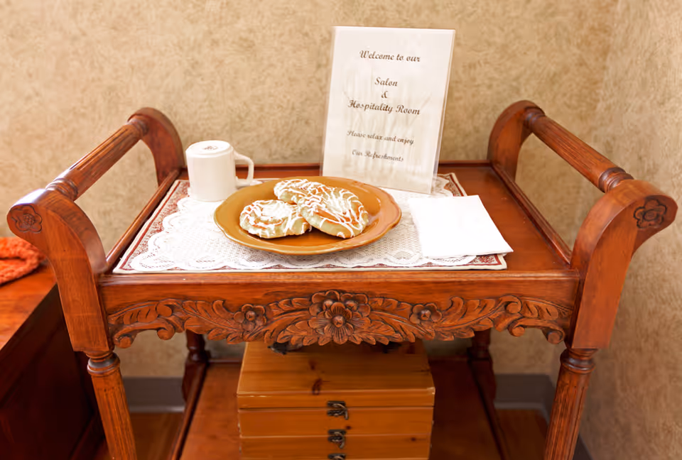 A wooden serving cart with carved floral designs holds a plate of three iced pastries, a white mug, a folded napkin, and a sign that reads 'Welcome to our Salon & Hospitality Room Please relax and enjoy our refreshments.' The cart is placed against a beige textured wall.