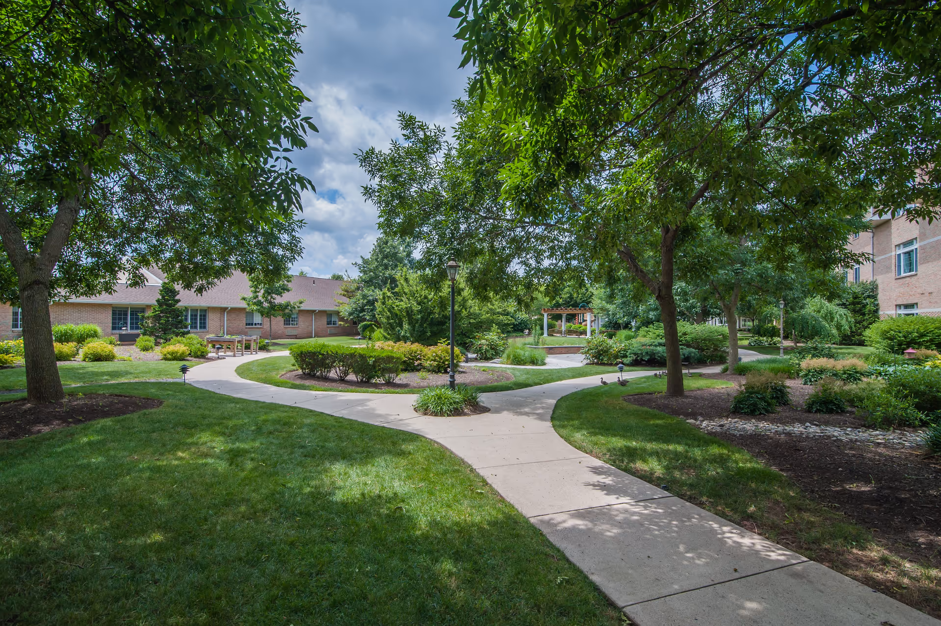 A peaceful outdoor garden area at Dock Woods – Living Branches Senior Living Community with a concrete pathway splitting into two directions, surrounded by green grass, trees, shrubs, and bushes. There are buildings in the background and a cloudy sky overhead.