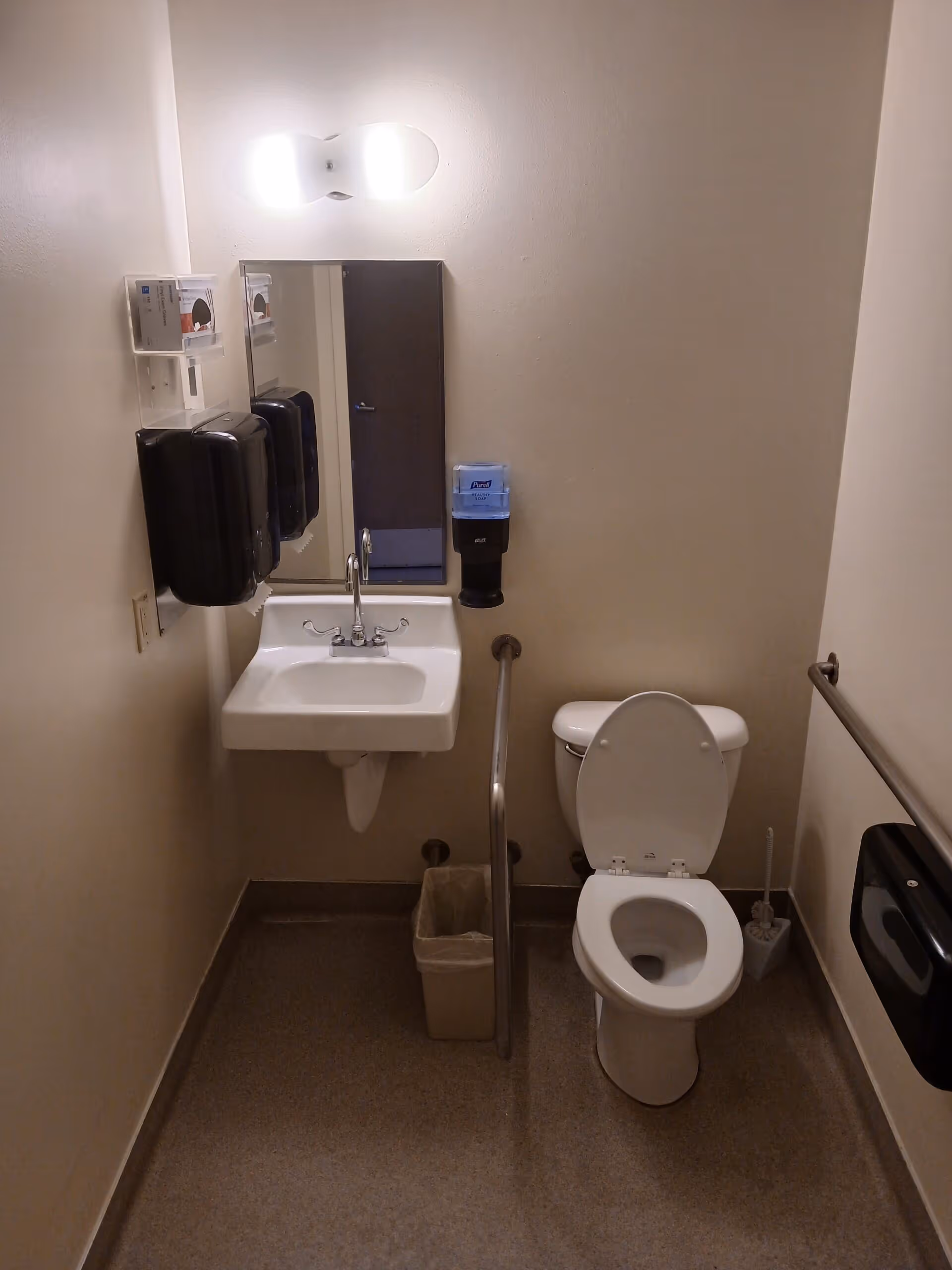 A small, clean bathroom with a white toilet, a white sink with a mirror above it, a trash bin, and handrails on both sides of the toilet. There are soap dispensers mounted on the wall near the sink and toilet. The walls are plain and light-colored, and the floor is covered with a speckled material.