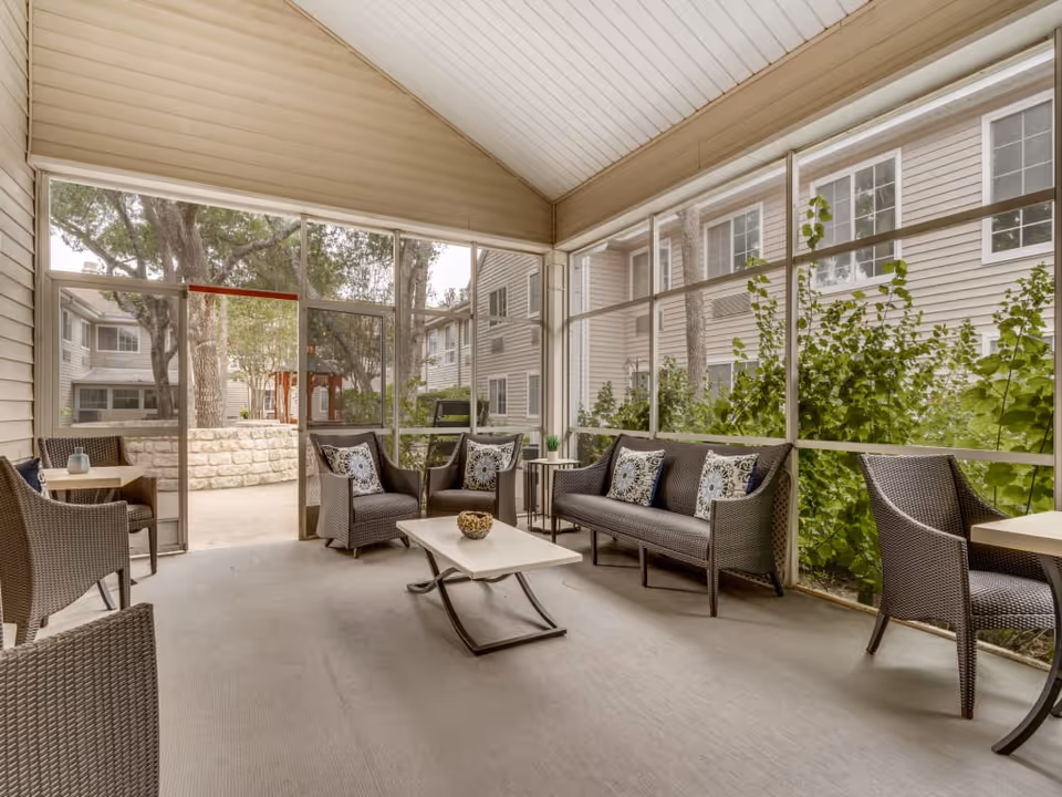 A screened-in porch area with wicker furniture including a sofa, armchairs, and small tables. The porch overlooks a courtyard with trees and shrubs, surrounded by a multi-story building with beige siding and white-framed windows.