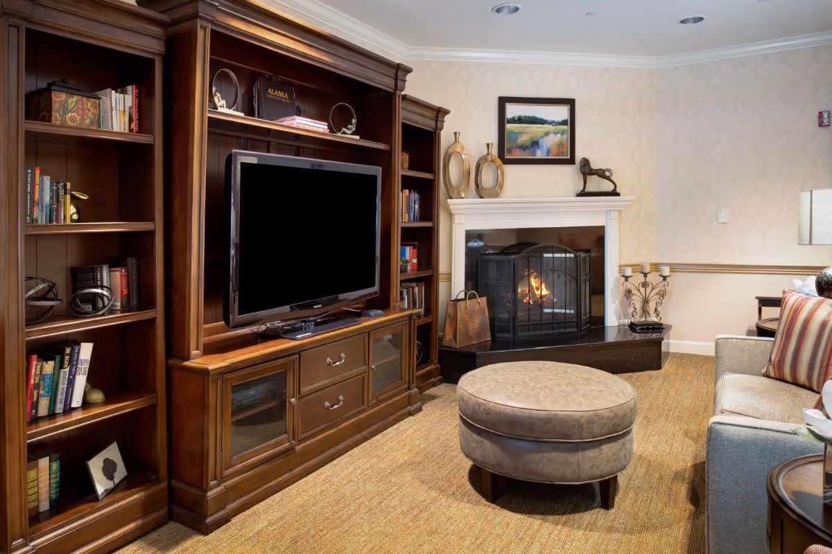 Living room with a large wooden entertainment center and TV, a lit fireplace, round ottoman, and sofa.