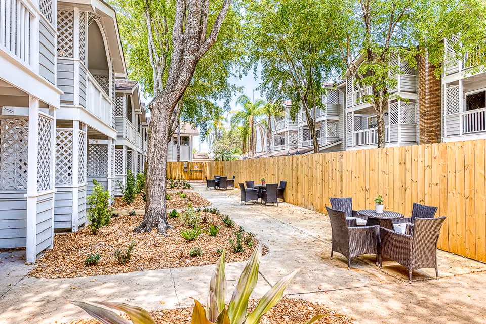 Outdoor patio area at Meridian at Lantana featuring several round tables with four wicker chairs each, surrounded by a wooden fence and landscaping with trees and plants. The patio is situated between two buildings with balconies and lattice railings.