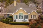 Single-story yellow cottage-style building with a large arched window, patio chairs and table, and surrounding shrubs and lawn.